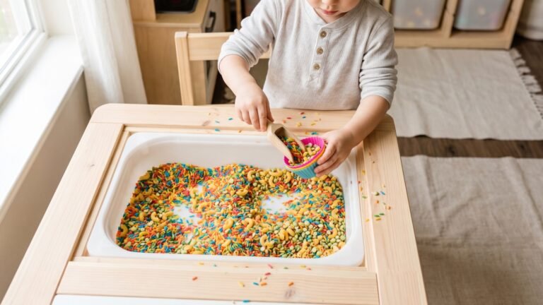 Toddler scooping colorful dyed rice and pasta into a silicone baking cup inside a wooden sensory table