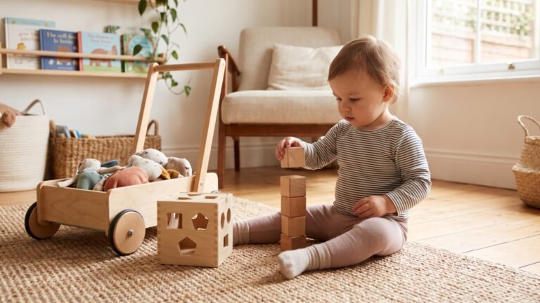 Toddler stacking wooden blocks on a rug beside a shape sorter cube and wooden wagon